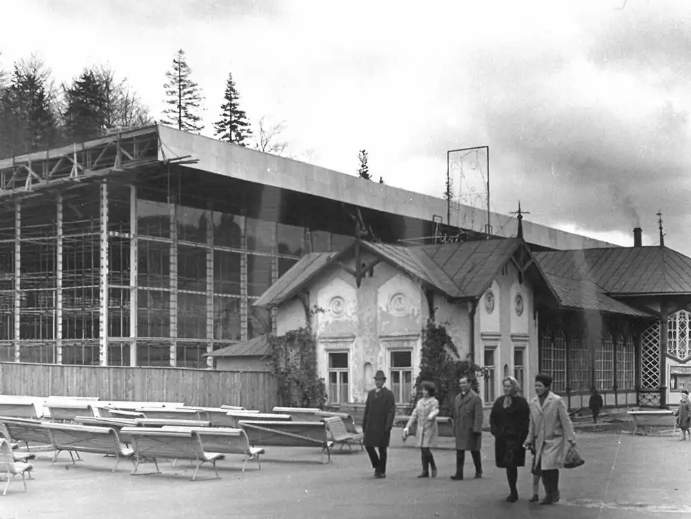 Archiwalne, czarno-białe zdjęcie budowy Pijalni Głównej w Krynicy-Zdroju — za rozbieraną, zdobioną drewnianą pijalnią wznosi się szkielet nowego, modernistycznego pawilonu z przeszkloną fasadą; przed budynkiem spacerują ludzie w płaszczach, obok rzędy złożonych ławek.