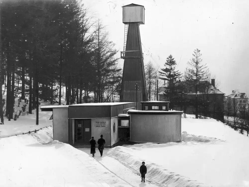 Archiwalne, czarno-białe zdjęcie odwiertu Zuber w Krynicy-Zdroju zimą — modernistyczny, biały pawilon o zaokrąglonej bryle stoi wśród zasp, nad nim drewniana wieża wiertnicza, w tle ośnieżony las i zabudowania uzdrowiska.