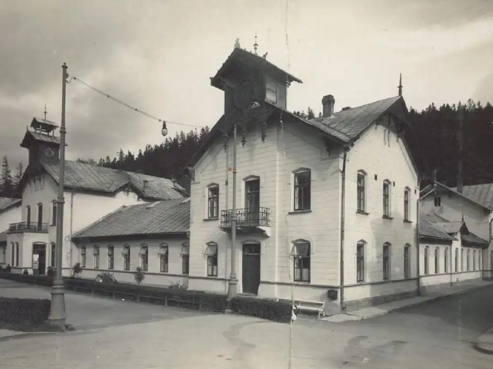 Archiwalne, czarno-białe zdjęcie Starych Łazienek Mineralnych w Krynicy-Zdroju — jasny, dwukondygnacyjny budynek w stylu uzdrowiskowym z wieżyczką i balkonem, po lewej niższe skrzydło z wieżą zegarową, w tle zalesione wzgórze.