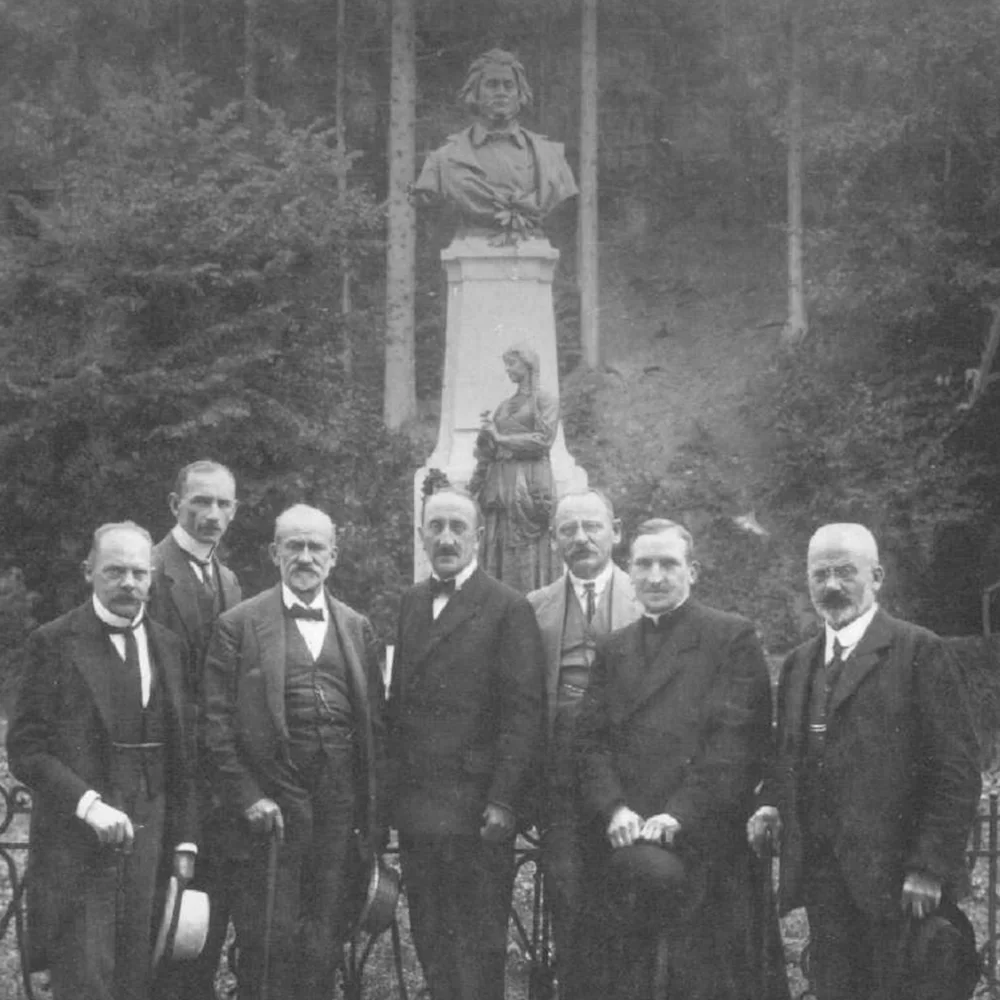 Czarno-białe, archiwalne zdjęcie przedstawiające 7 mężczyzn, elegancko ubranych i trzymających w rękach kapelusze na tle pomnika.