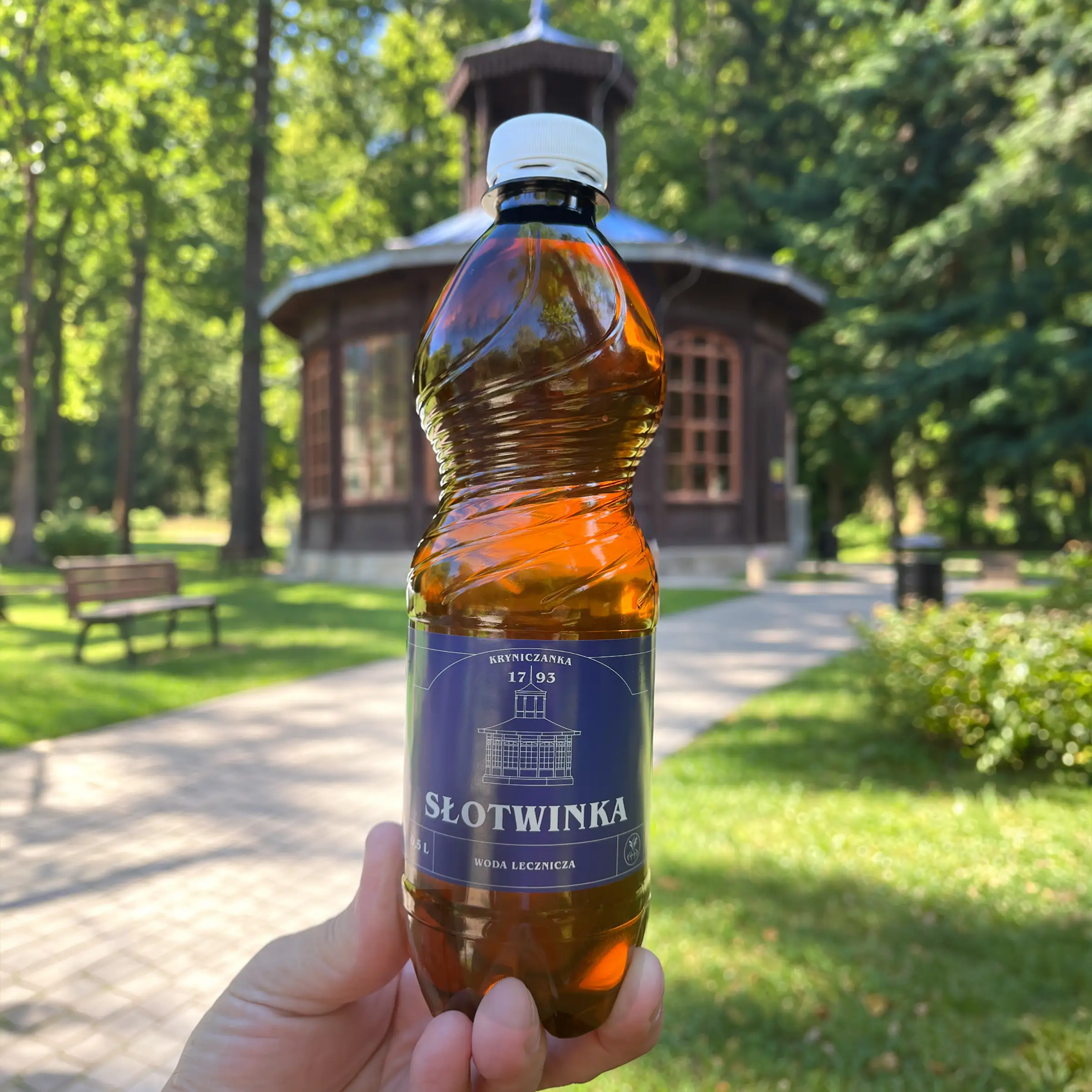 Dłoń trzymająca butelkę wody leczniczej Słotwinka 0,5 l na tle drewnianego pawilonu Pijalni Słotwinka w Krynicy-Zdroju, otoczonego zielenią.